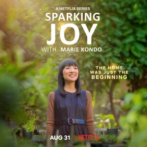 Sparking Joy with Marie Kondo. Marie Kondo ist vor Bäumen und Sträuchern abgebildet. Sie lächelt.