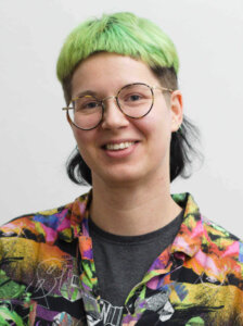 Elis Eder im Porträt. Elis ist weiß, hat kurze, grüne Haare und trägt eine Brille und ein buntes Hemd.