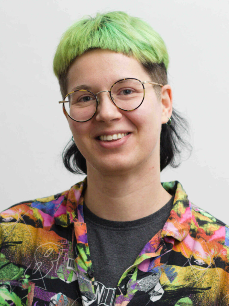 Das bin ich: Elis Eder. Elis ist eine weiße Person. Elis hat kurze, grüne Haare, trägt eine Brille und ein buntes Hemd. Elis lächelt in die Kamera.